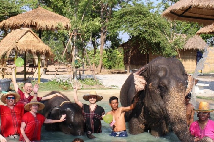 Group of people and elephants playing in water under a sunny sky at an elephant park.