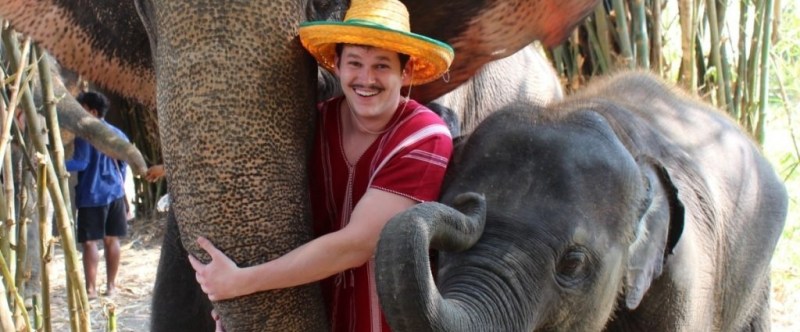 Person in a straw hat hugs an adult elephant with a baby elephant nearby among trees.