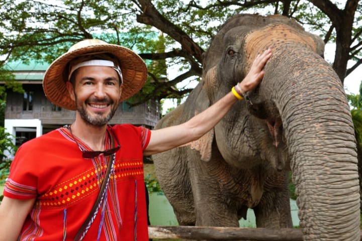 Man in red shirt and straw hat smiling, standing next to an elephant in a park setting.
