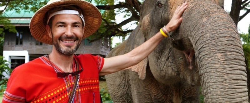 Man in red shirt and straw hat smiling, standing next to an elephant in a park setting.