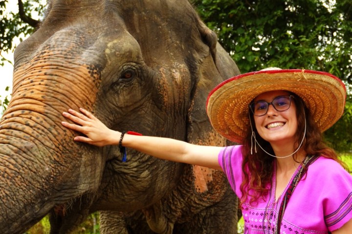 Woman in pink dress and straw hat smiling and touching an elephant's head outdoors.