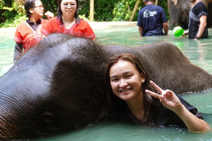 Smiling person with elephant in water, making a peace sign with hand.