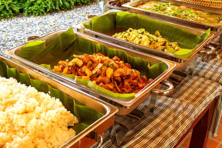 Buffet table with trays of rice and various dishes, lined with banana leaves, set outdoors.