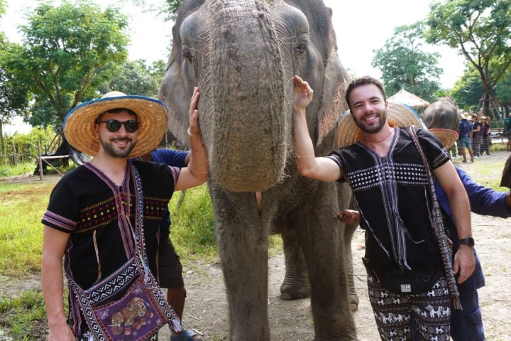 Two people in patterned clothes smiling with an elephant outdoors.