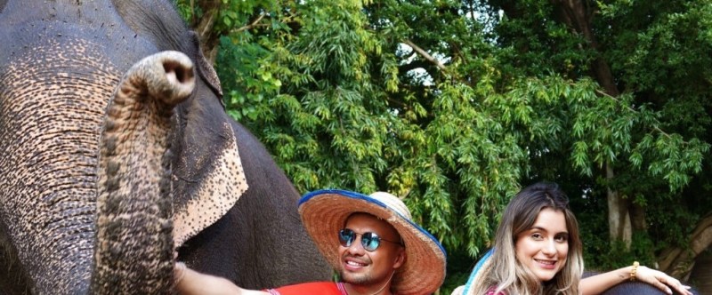 Two people smiling, standing next to an elephant in front of lush green trees.