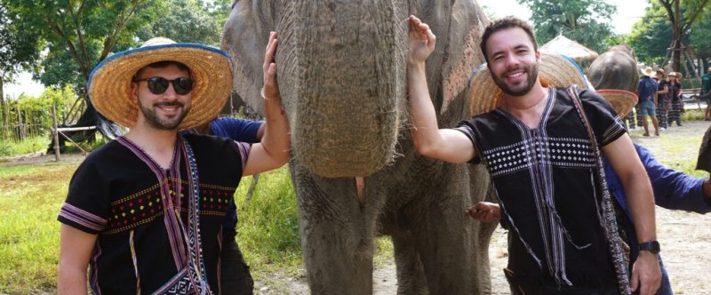Two men in traditional attire smiling and touching an elephant's trunk outdoors.