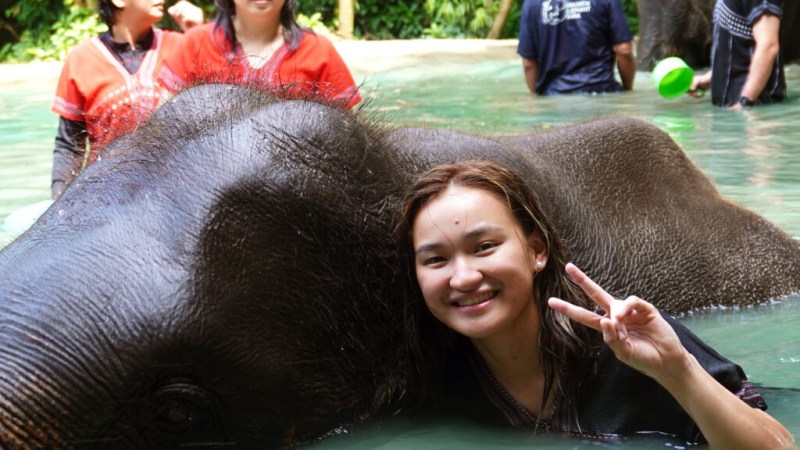 Woman smiling with peace sign next to an elephant in water, others in background.