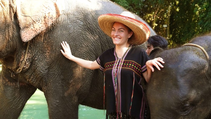 Person in a straw hat smiling, standing between two elephants in an outdoor setting.