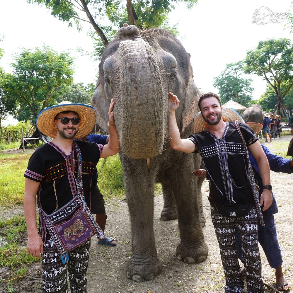 Two smiling men in patterned outfits pose with an elephant outdoors on a sunny day.