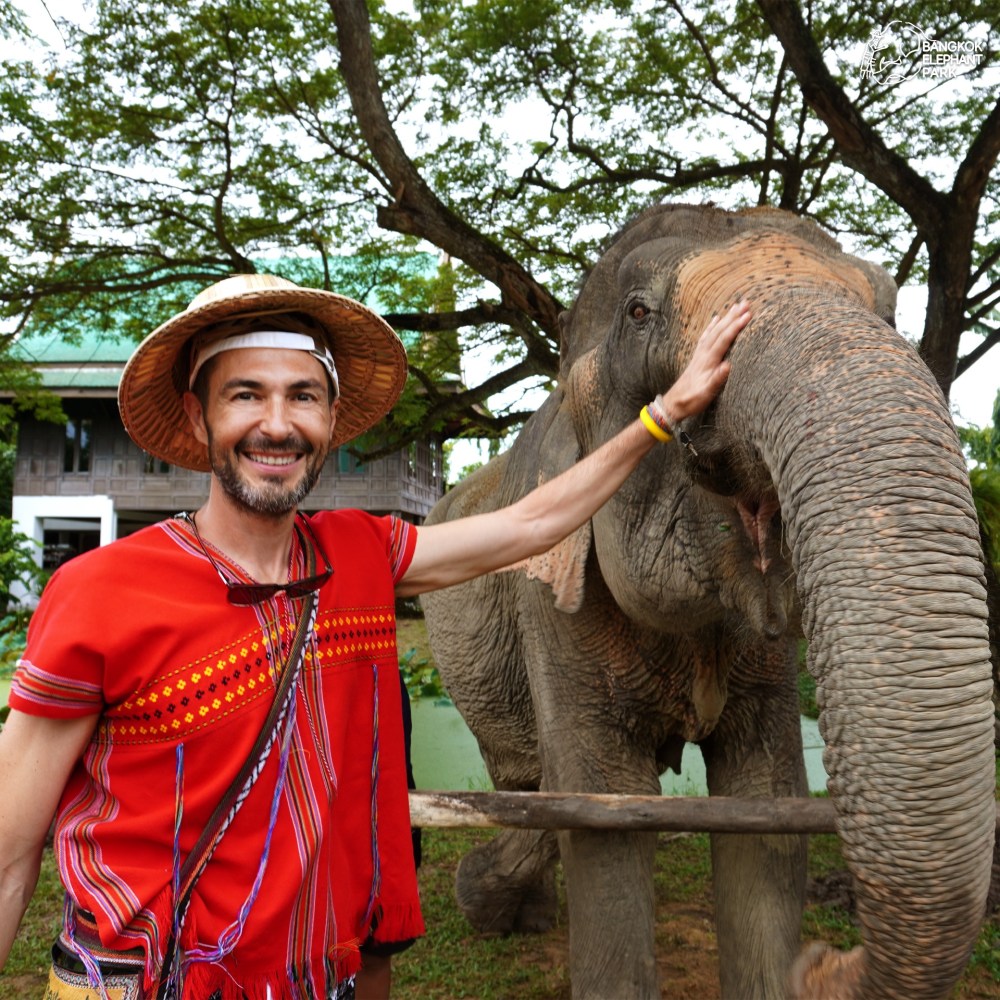 Man in red attire and straw hat smiling while touching elephant's trunk outdoors.