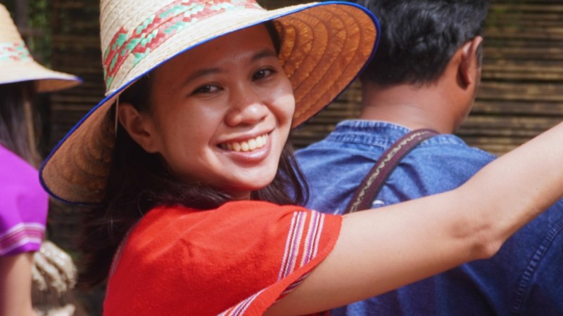 Smiling person in a traditional hat and red patterned shirt, holding mud at an outdoor event.