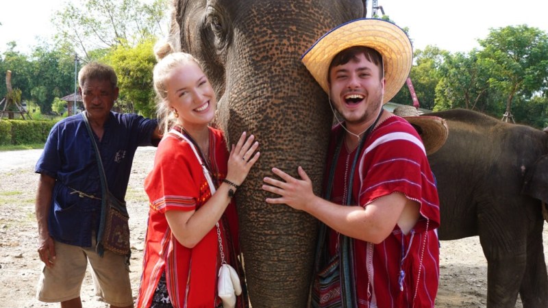 Two people smiling and hugging an elephant, wearing colorful clothing outdoors.
