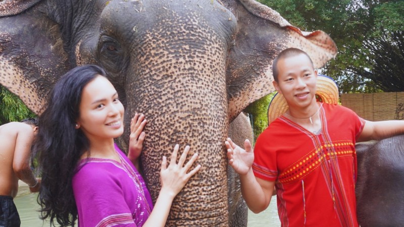 Two people in colorful attire stand beside an elephant in an outdoor setting.