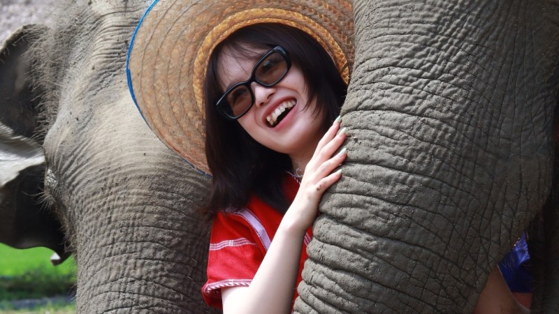Person in hat and glasses smiling, embraced by an elephant's trunk.